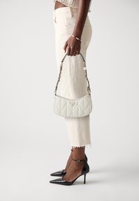 Quilted white handbag with a short chain strap, featuring a metal logo emblem. Model wears light beige jeans and black pointed heels.