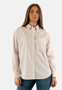 Camisa de botones a rayas en beige y blanco, con un bolsillo en el pecho, cuello con puntas y mangas largas con puños abotonados.