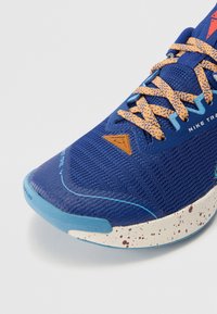 Chaussure de sport bleue avec une tige en tissu texturé, semelle parsemée d'orange et accents bleus contrastants. Caractéristiques : lacets plats et logo triangulaire.