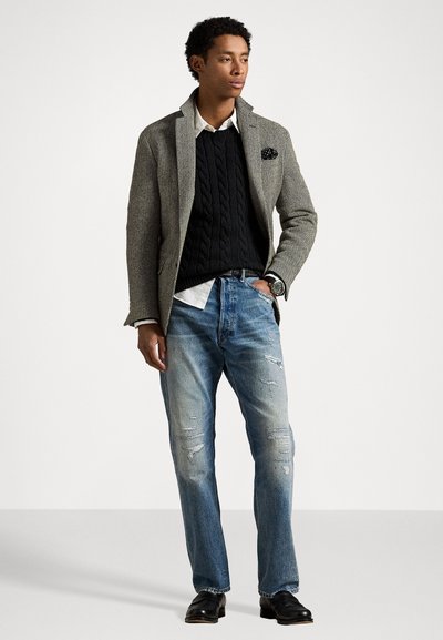 Blazer en pied-de-poule gris sur un pull en laine noire à mailles torsadées, chemise blanche, jeans bleus déchirés et chaussures noires. Complété par un mouchoir de poche.