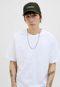 Witte T-shirt met korte mouwen, gedragen met een zilveren kettinghanger; olijfgroene pet met het "JJ Studios" logo in het wit. Lichte achtergrond.