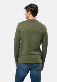 Chemise à manches longues de couleur olive, fabriquée en matériau doux, avec un col rond et un design simple, sans ornements. Portée avec un jean en denim bleu.