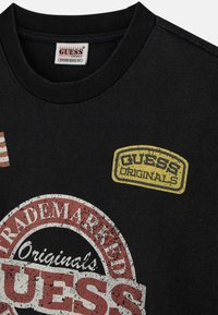 Zwarte t-shirt met verwoeste grafische prints, waaronder een ronde logo in wit en rood, en een gele "GUESS ORIGINALS" patch.