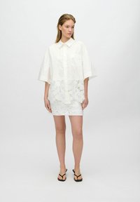 Robe blanche avec un col, des manches courtes et un ourlet en dentelle à motif floral, associée à des tongs noires.