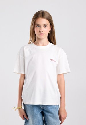 Jonge vrouw met lang bruin haar draagt een wit T-shirt met korte mouwen en een blauwe spijkerbroek, staand tegen een effen lichte achtergrond.