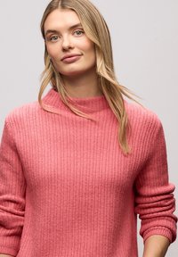 Pull côtelé rose avec un col montant et des poignets retroussés, fabriqué en tissu texturé. La couleur est un rose doux et chaleureux.