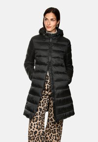 Schwarze Daunenjacke mit Kapuze, mit gesteppten Abschnitten und einem Reißverschluss auf der Vorderseite. Getragen über Leoprint-Hosen. Glatte Textur.