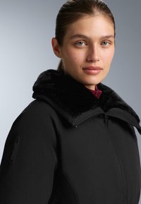 Crna jakna s velikim, mekanim ovratnikom od umjetnog krzna, s prednjim patentnim zatvaračem i RECCO oznakom na rukavu. Glatka tekstura i kroj prilagođen tijelu.