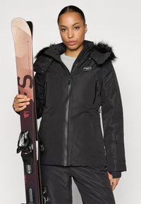 Μαύρο μπουφάν σκι με κουκούλα επενδεδυμένη με γούνα, τσέπες με φερμουάρ και λείο υλικό. Το �μοντέλο κρατάει σκι με κόκκινες και μαύρες λεπτομέρειες.