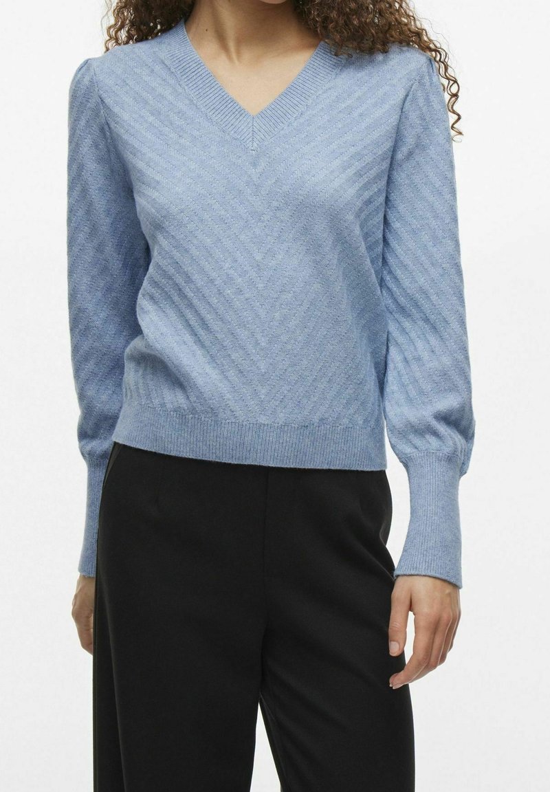 Pull en tricot bleu clair avec un décolleté en V, présentant un motif diagonal, des poignets côtelés et une coupe légèrement cropped. Associé à un pantalon noir.