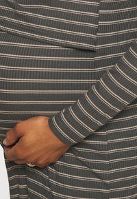 Robe en tissu côtelé gris avec des rayures verticales crème ; manches longues et coupe ajustée. Gros plan sur une main reposant sur la taille.