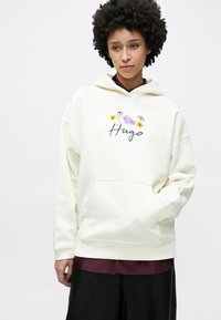 Krém kapucnis pulóver elülső zsebbel, virágmintával és a "Hug" szóval díszítve, írott betűtípussal. Puha anyagból, laza szabású és állítható kapucnival.