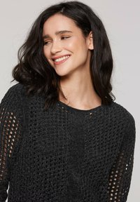 Junge Frau mit schulterlangem, dunklem Haar, die einen strukturierten, schwarzen Strickpullover trägt, lächelt und nach links schaut.