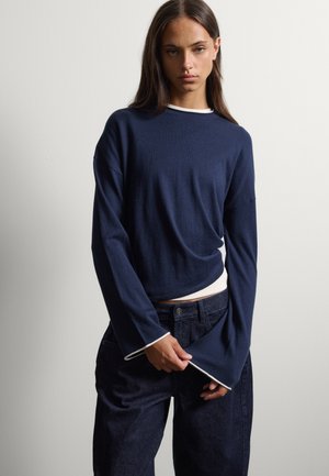 Vrouw met lang donker haar die een marineblauwe trui met lange mouwen en witte bies en donkere spijkerbroek draagt tegen een effen lichte achtergrond.