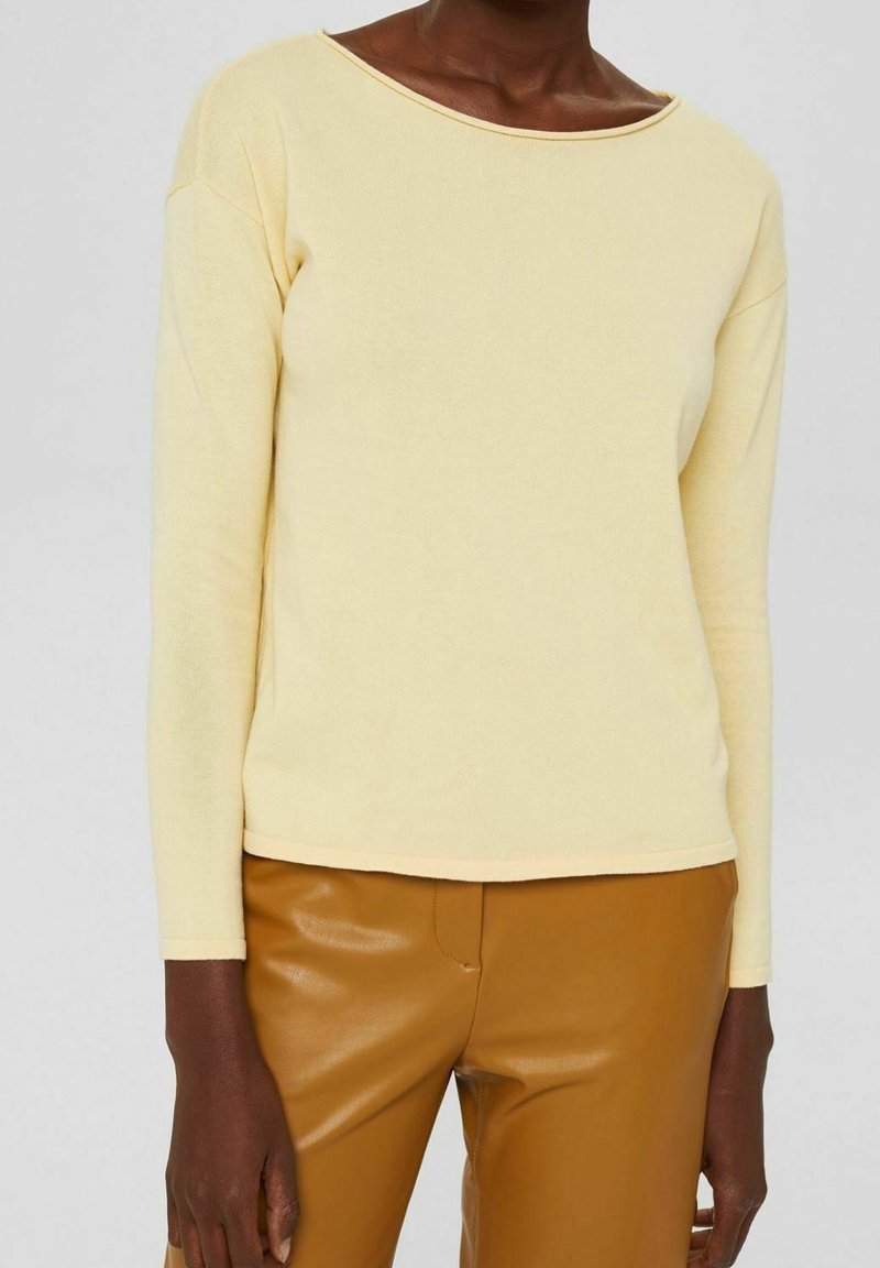 Personne portant un haut à manches longues jaune pâle avec un large col et un pantalon en cuir marron, visible du cou aux hanches.