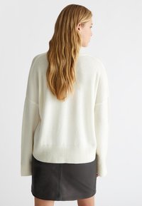 Maglione oversize color crema con orlo a coste, caratterizzato da una consistenza morbida. Abbinato a una mini gonna in pelle nera. Vista da dietro.