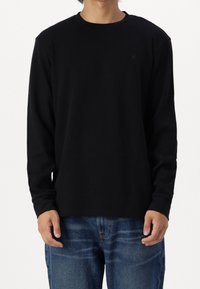 Maglietta nera a maniche lunghe in tessuto a costine, con scollatura a girocollo e un discreto logo sulla parte anteriore, abbinata a jeans blu.