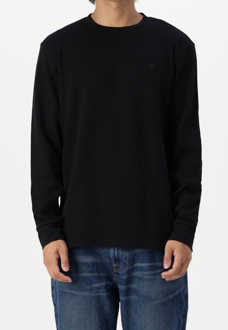 Maglietta nera a maniche lunghe in tessuto a costine, con scollatura a girocollo e un discreto logo sulla parte anteriore, abbinata a jeans blu.