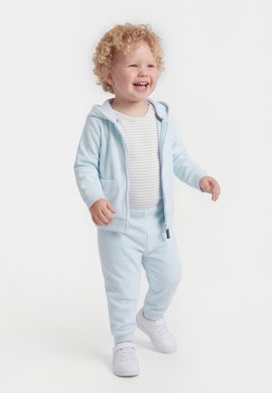 Toddler with curly blond hair smiling, wearing light blue hoodie, striped shirt, matching pants, and white sneakers, standing on white background.