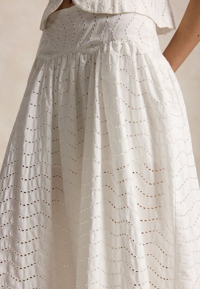 Λευκό φόρεμα από ύφασμα με λευκές τρύπες, με κυματιστά σχέδια κοπής, με σούρα στη μέση και πλαϊνή τσέπη στην οποία ακουμπάει ένα χέρι.