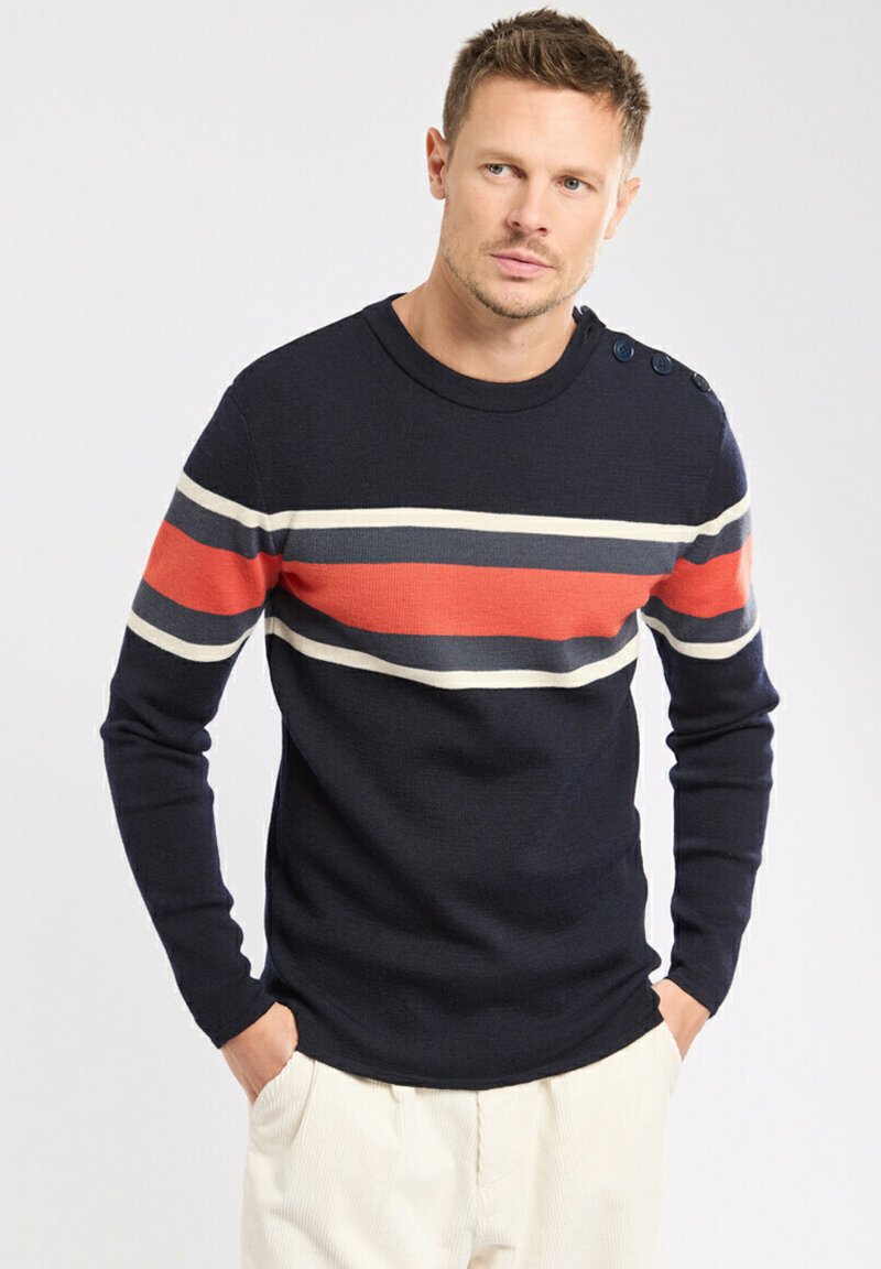Homme portant un pull marine avec des rayures horizontales blanches, gris foncé et rouges, et un pantalon beige, se tenant les mains dans les poches.
