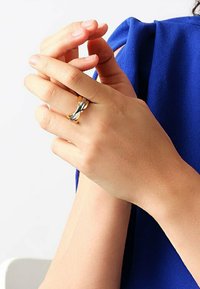 Guld- och silverring i en fläta på en hand, som visar en slät, polerad yta och en modern, flytande design mot en blå tygbakgrund.