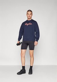 Férfi, aki sötétkék Rapha pulóvert, fekete kerékpáros rövidnadrágot, fekete zoknit és sportcipőt visel, fekete vízflakont tart, és sima háttér előtt áll.