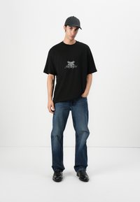 Black oversized t-shirt with a graphic design at the center, paired with blue distressed jeans and black shoes, worn with a black cap.