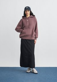 Νεαρή γυναίκα που φοράει μωβ φούτερ Nike, μαύρη μακριά φούστα, λευκά αθλητικά παπούτσια και μαύρο καπέλο Nike, στέκεται σε μπλε χαλί μπροστά σε λευκό τοίχο.