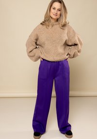 Beige gebreide trui met hoge hals en ballonmouwen, gecombineerd met losse paarse broek en zwarte instappers. Glad, gestructureerd materiaal.