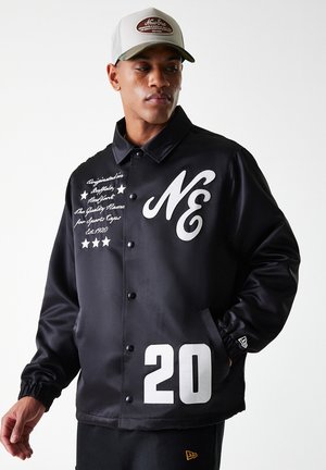 Veste en satin noir avec logo "NE" blanc, texte brodé et le numéro "20" sur le bas. Comprend une fermeture à boutons-pression et des poches.