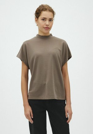 Junge Frau mit lockigem Haar, die ein braunes Kurzarmoberteil und schwarze Hose trägt, steht vor einem schlichten weißen Hintergrund.