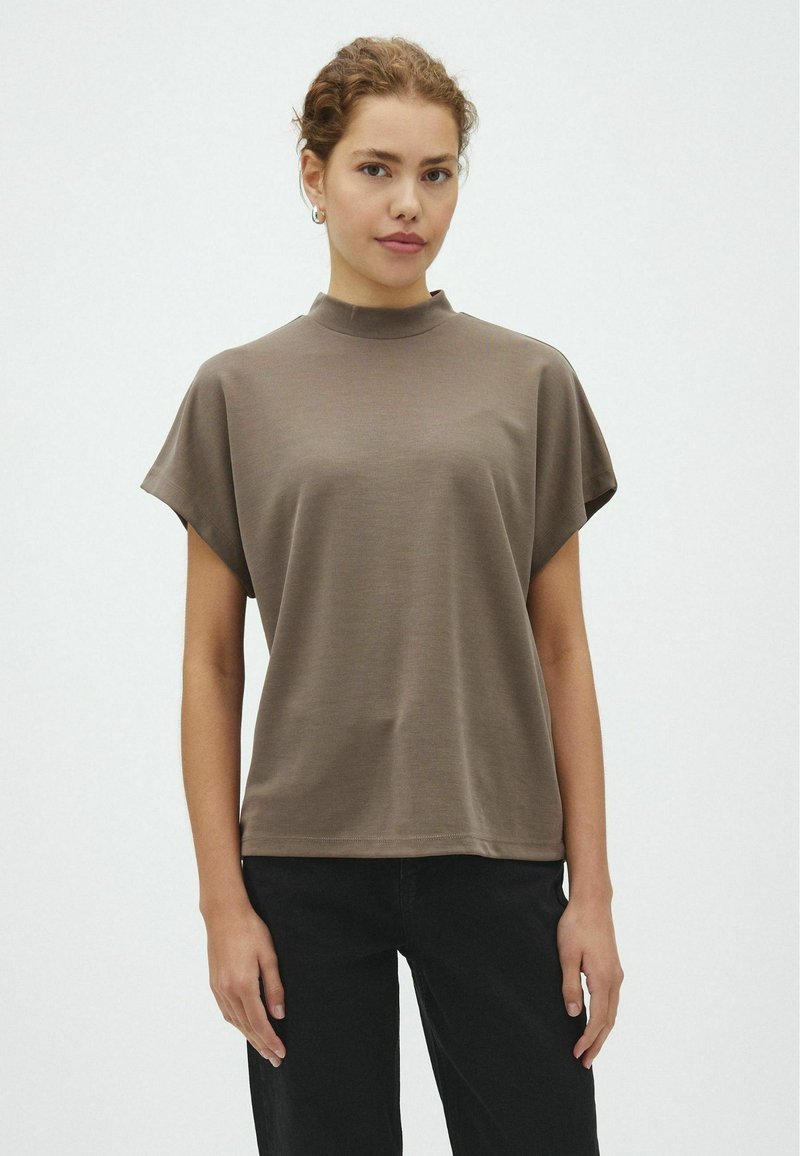 Jeune femme aux cheveux bouclés portant un haut marron à manches courtes et un pantalon noir, debout devant un fond blanc uni.
