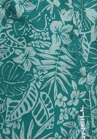 Tyrkysová látka s jemnými botanickými vzormi obsahujúcimi listy a kvety; na boku je biely značkový nápis. Hladká textúra.