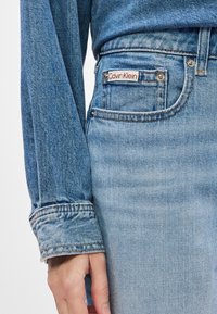 Svetlo modri kavbojke iz denima s klasičnim pet-pocket dizajnom. Imajo rdečo in belo oznako Calvin Klein na zadnjem žepu.