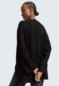 Vrouw met gevlochten haar in een lage knot, gekleed in een losse zwarte langemouwen top, staande met haar handen achter haar rug samengevoegd.
