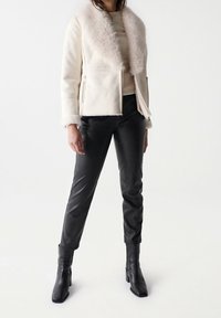 Cremefarbener Schafsfelljacke mit einem Pelzkragen, kombiniert mit schwarzen Kunstlederhosen und schwarzen Ankle Boots mit Absatz. Einfaches, maßgeschneidertes Design.