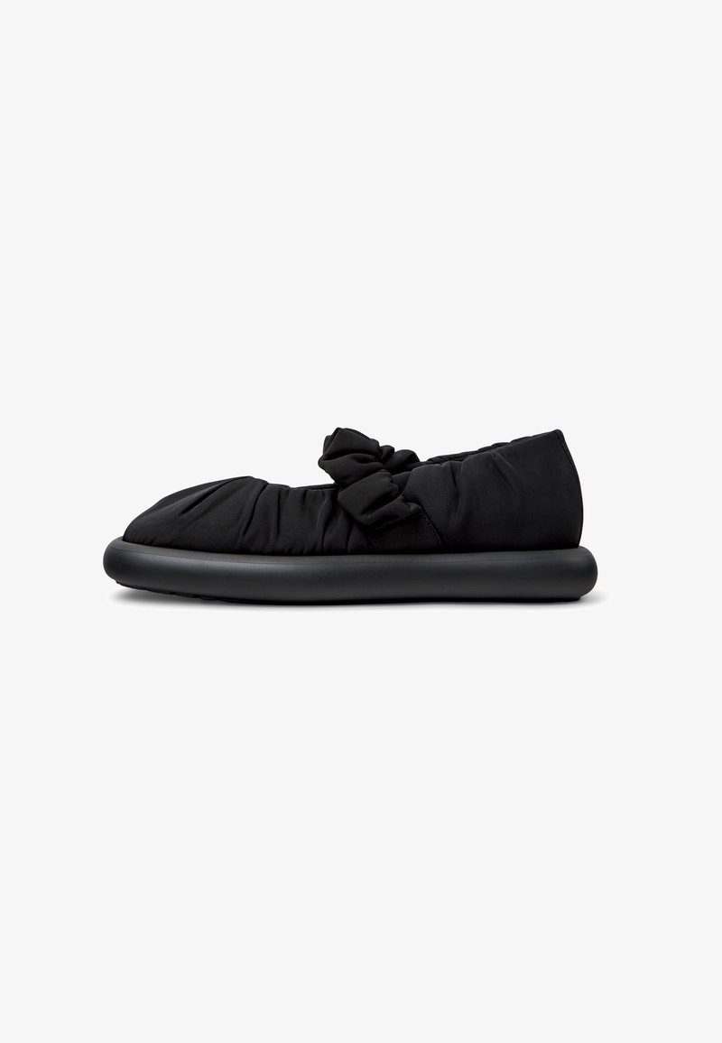 Zapato negro tipo slip-on con una parte superior de textil suave y recogida, adornado con un detalle de lazo, que cuenta con una suela de goma lisa y redondeada.