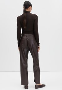 Woman with long dark hair in a ponytail wears dark brown turtleneck, high-waisted faux leather pants, and matching flat shoes, standing facing white wall.