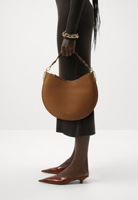 Brown leather handbag in a crescent shape, featuring a textured surface and gold hardware. Worn with a fitted ribbed brown dress and pointed heels.
