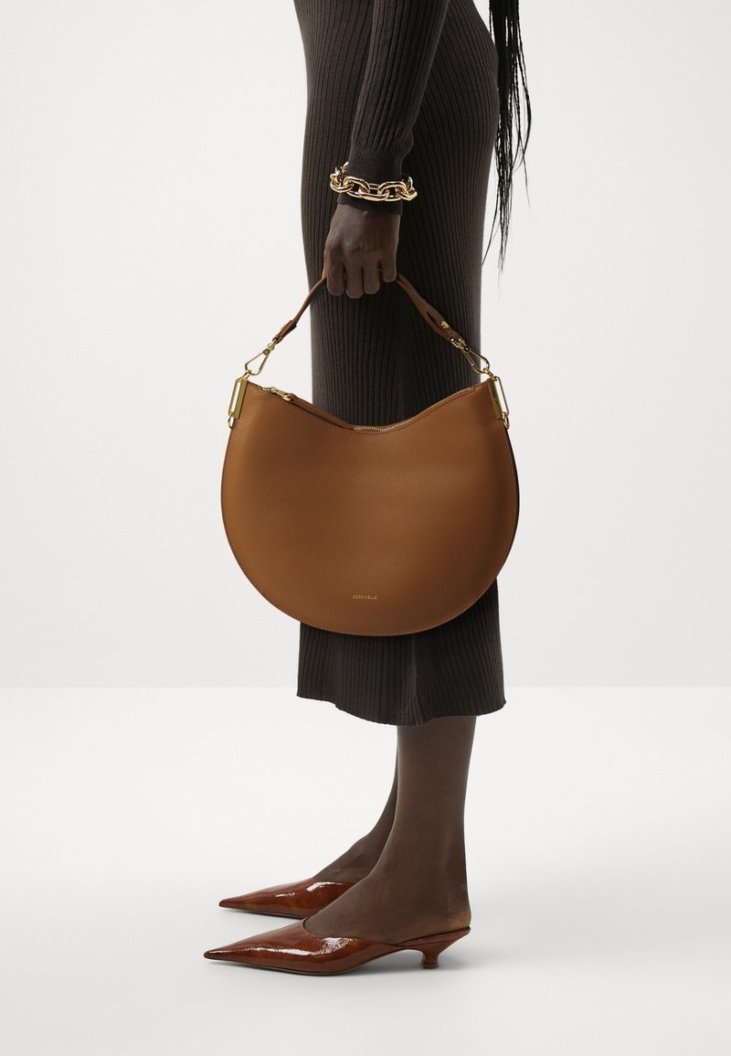 Brown leather handbag in a crescent shape, featuring a textured surface and gold hardware. Worn with a fitted ribbed brown dress and pointed heels.