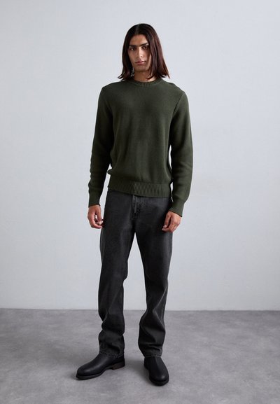Dunkelgrüner Strickpullover mit geripptem Saum, kombiniert mit dunkelgrauen Jeans und schwarzen Stiefeln mit Knöchelschnitt. Das Model steht vor einem hellgrauen Hintergrund.