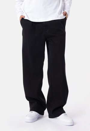 Personne portant un pantalon noir ample, des baskets blanches et une chemise blanche à manches longues, les mains dans les poches sur un fond blanc.