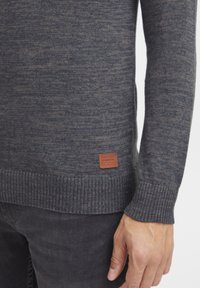 Nahaufnahme einer Person, die einen grauen Strickpullover mit einem braunen Leder-Brandpatch in der Nähe des Saums und dunkelgraue Hosen trägt.