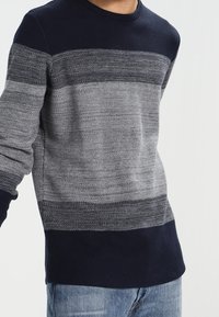 Man med en långärmad stickad tröja i horisontella grå och marinblå ränder, kombinerad med ljusblå jeans.