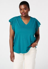 T-shirt en coton teal avec un col en V et des manches à dentelle. Texture douce, coupe décontractée, associé à un pantalon clair.