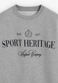 Sudadera gris hecha de una mezcla de algodón. Presenta un bold texto negro "SPORT HERITAGE" y un texto más pequeño "Scalpers Company" con un año de fundación y acentos de logo.