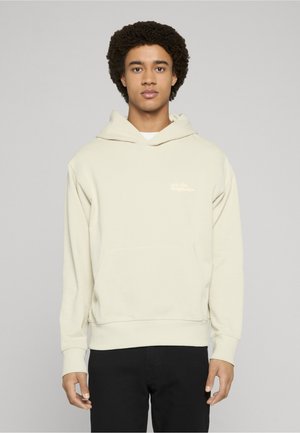 Noor mees lokkis juustega, kandes helebeeži kapuutsiga pusa ja musti pükse, seisab halli ühevärvilise tausta ees.