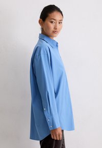 Jonge vrouw met donker haar dat naar achteren is vastgebonden, gekleed in een oversized lichtblauw overhemd met knopen, staand in profiel tegen een simpele witte muur.