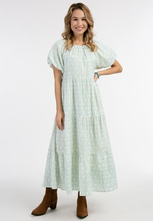 Lichtgroene maxi-jurk met bloemenpatroon, pofmouwen, gelaagd ontwerp en een ontspannen pasvorm, gecombineerd met bruine enkellaarsjes.